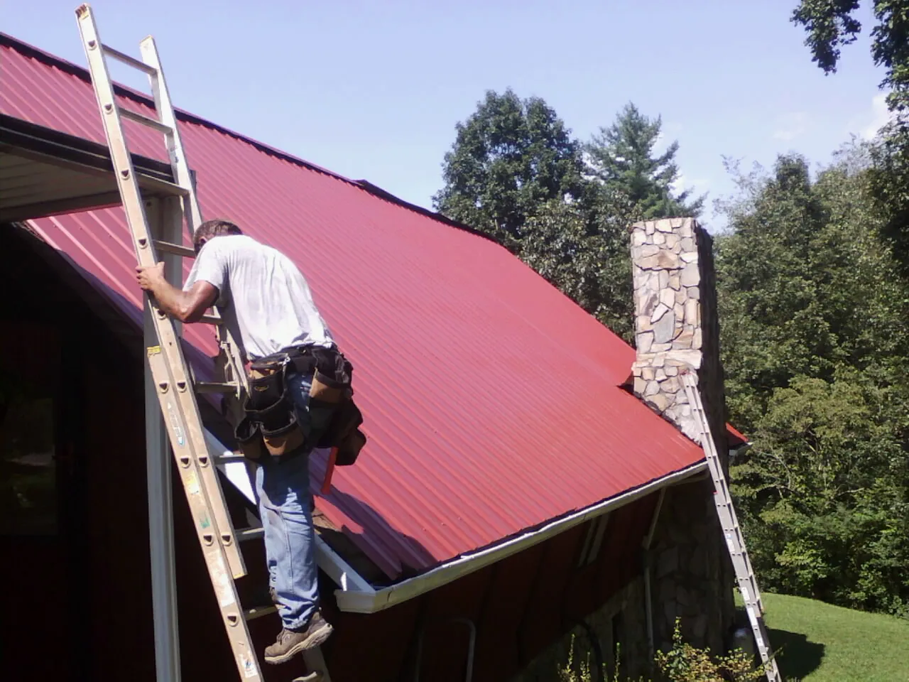 Roofer on a ladder beside a red metal roof and stone chimney — Soffit & Fascia Repair by Roofing Brothers in Fort Howard